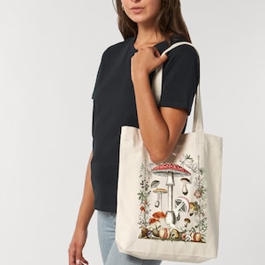 May include: A white canvas tote bag with a colorful illustration of mushrooms. The illustration features a large red and white mushroom with smaller mushrooms surrounding it.