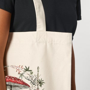 May include: A white canvas tote bag with a floral and mushroom design. The bag has a long white strap and features a red and white mushroom with a white stem.