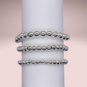 May include: Three silver bead bracelets stacked on a white surface. The bracelets are made of small, round beads.