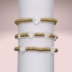 May include: Three gold beaded bracelets with white shell charms in the shape of a four-leaf clover. The bracelets are stacked on a white display stand.
