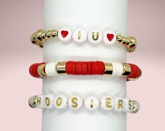 May include: A set of three beaded bracelets. The top bracelet features white beads with red hearts and the letters I and U in gold. The middle bracelet has red and white disc beads with gold spacers. The bottom bracelet has white beads with the gold text HOOSIERS.