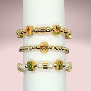 May include: Three stretch bracelets featuring gold-toned metallic beads and white pearl-like beads. Each bracelet incorporates rectangular charms decorated with colorful Mahjong tile symbols, including red, green, and black characters and patterns.