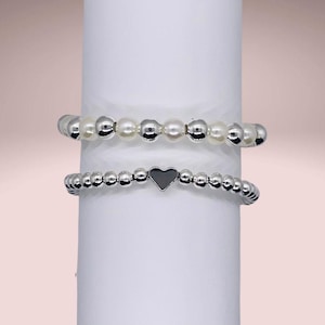 May include: Two silver beaded bracelets, one with white pearls and the other with a silver heart charm.