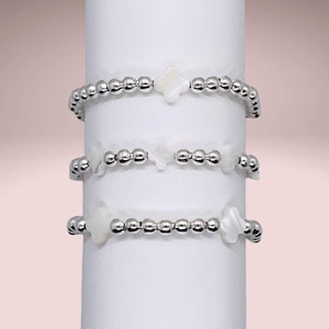 May include: Three silver beaded bracelets with white shell charms in the shape of a four-leaf clover.