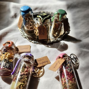 Peut inclure: Cinq petits flacons en verre contenant diverses herbes et fleurs. Chaque flacon est scellé par une cire de couleur différente et porte une étiquette avec un texte manuscrit. Les étiquettes indiquent "Glamour", "Renforcement psychique", "Amour", "Bouclier énergétique" et "Protection".