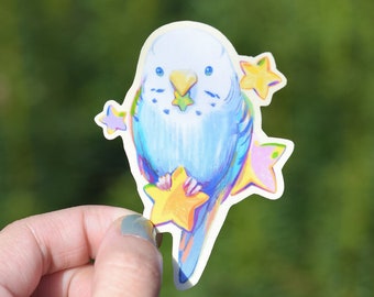 May include: A blue and yellow sticker featuring a cartoon blue budgie perched on a yellow star. The budgie is surrounded by smaller stars in pink, yellow, and purple.