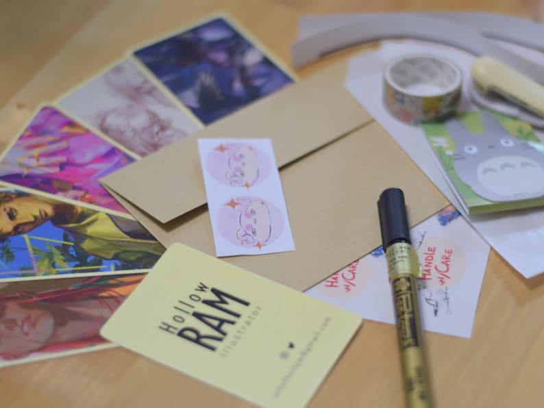 Puede incluir: Un surtido de impresiones art&iacute;sticas, pegatinas y art&iacute;culos de papeler&iacute;a. Incluye una tarjeta de visita con el texto "Hollow RAM Illustrator", cinta adhesiva con estampado floral y un rotulador dorado. Los art&iacute;culos est&aacute;n dispuestos sobre una superficie clara.