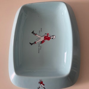 May include: A light blue, rectangular ashtray with rounded edges. It features a graphic of a man in a red coat and the text "BORN 1820 - STILL GOING STRONG". A smaller version of the graphic is at the bottom.