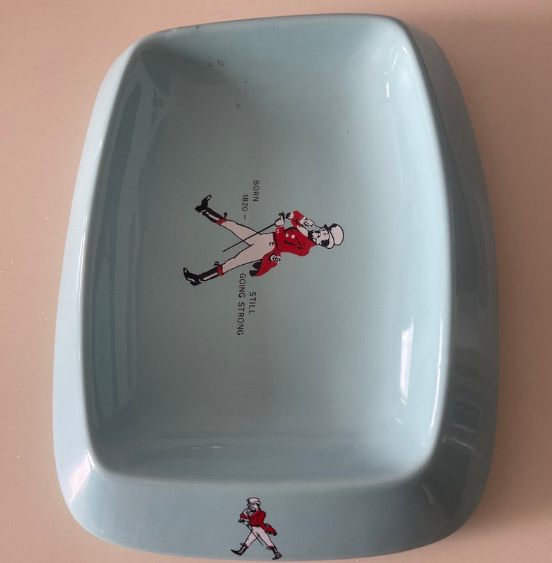 May include: A light blue, rectangular ashtray with rounded edges. It features a graphic of a man in a red coat and the text "BORN 1820 - STILL GOING STRONG". A smaller version of the graphic is at the bottom.