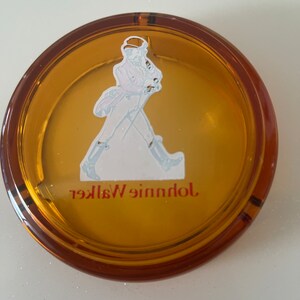 May include: Round amber-colored ashtray with a brown rim. The ashtray features a white and blue illustration of a man walking, with the words "Johnnie Walker" in red lettering. The ashtray is made of glass.