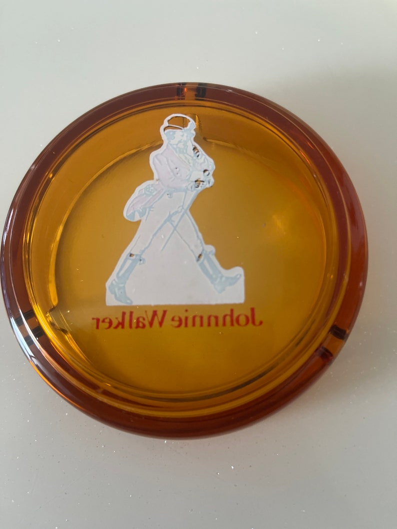 May include: Round amber-colored ashtray with a brown rim. The ashtray features a white and blue illustration of a man walking, with the words "Johnnie Walker" in red lettering. The ashtray is made of glass.