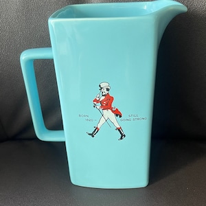 Op de afbeelding: Een lichtblauwe keramische kan met het Johnnie Walker-logo. Het logo toont een man in een rood jasje en witte broek die met een stok loopt. De tekst op het logo luidt "Born 1820 - Still Going Strong".