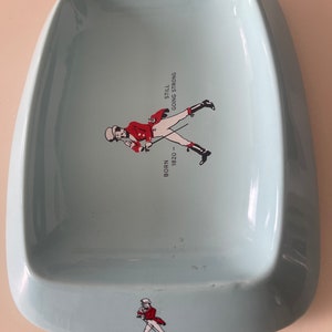 May include: A light blue, ceramic ashtray with a rounded rectangular shape. It features a graphic of a man in a red coat and the text "BORN 1820 - STILL GOING STRONG". A smaller version of the graphic is at the bottom.