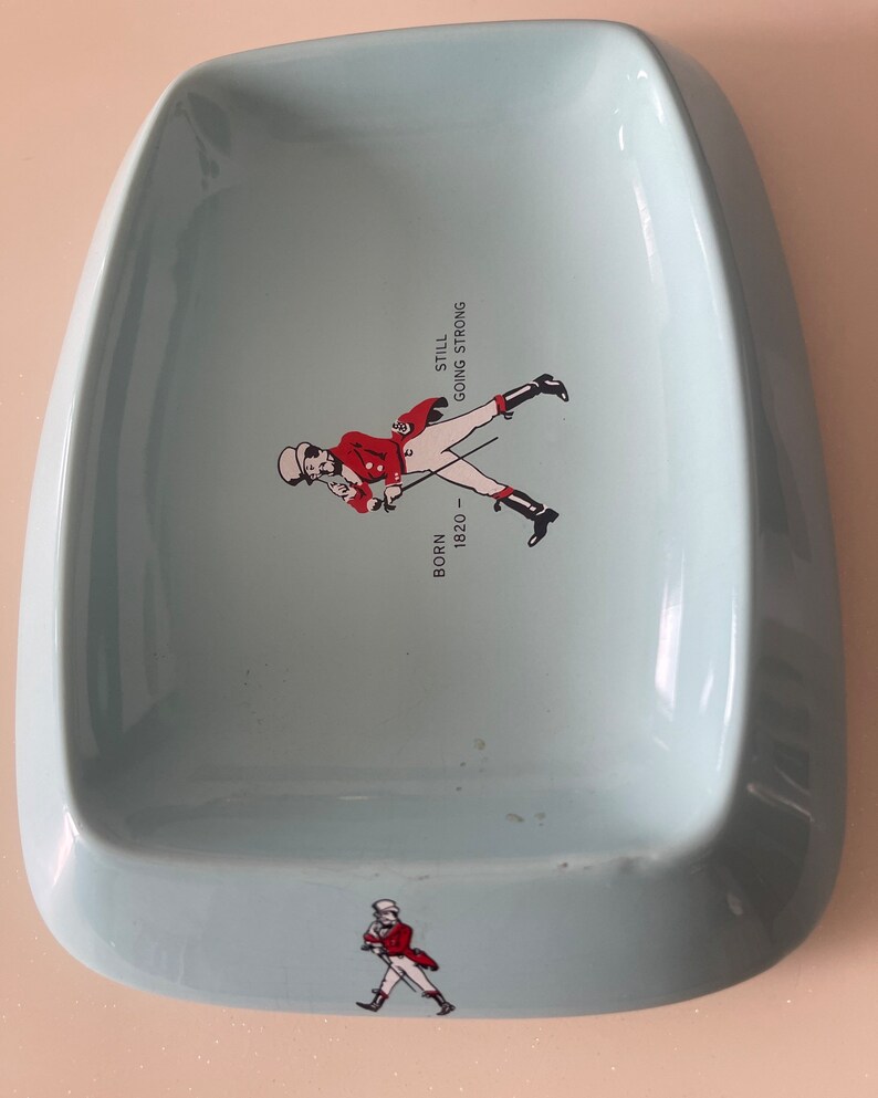 May include: A light blue, ceramic ashtray with a rounded rectangular shape. It features a graphic of a man in a red coat and the text "BORN 1820 - STILL GOING STRONG". A smaller version of the graphic is at the bottom.
