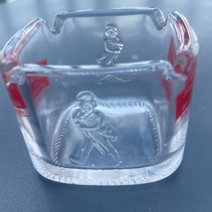 May include: A clear glass ashtray with a red and white label and a raised design of a person holding a bottle. The label has the text "Johnnie Walker" on it.