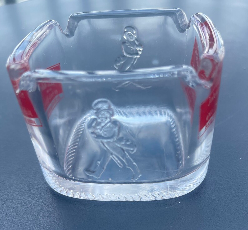 May include: A clear glass ashtray with a red and white label and a raised design of a person holding a bottle. The label has the text "Johnnie Walker" on it.