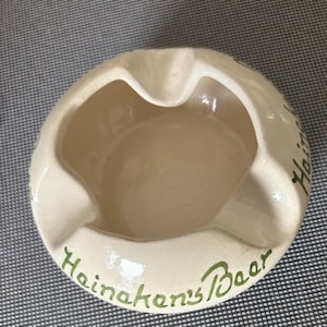 May include: A white ceramic ashtray with green lettering that reads "Heineken's Beer".