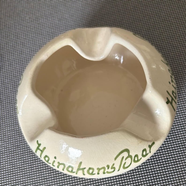 May include: A white ceramic ashtray with green lettering that reads "Heineken's Beer".