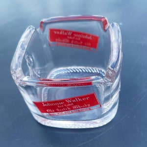 May include: A clear glass ashtray with a red Johnnie Walker logo. The logo reads "Johnnie Walker Red Label Old Scotch Whisky Born 1820 still going strong."