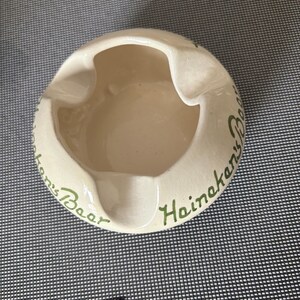 May include: White ceramic ashtray with green lettering that reads "Heineken's Beer".