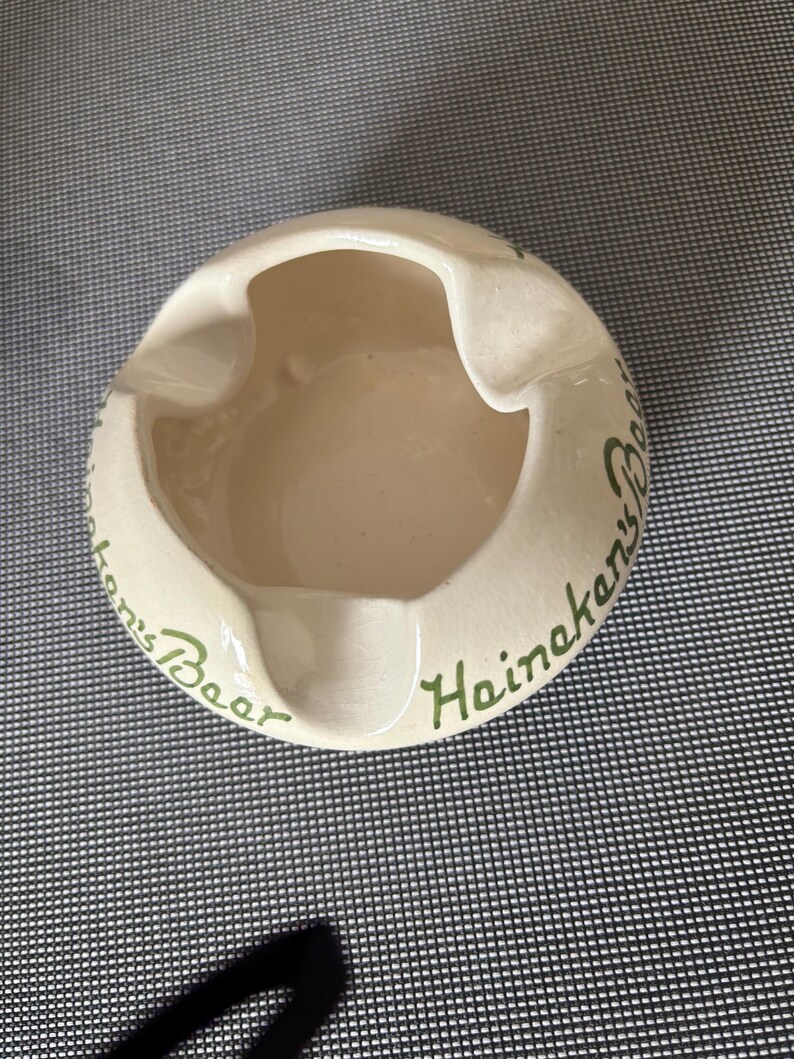 May include: White ceramic ashtray with green lettering that reads "Heineken's Beer".