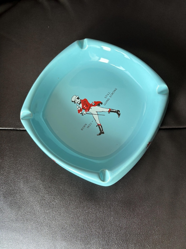 May include: A light blue ceramic ashtray with a white outline and a red and white illustration of a man in a red jacket and white trousers walking with a cane. The text "BORN 1820" and "STILL GOING STRONG" is printed on the ashtray.