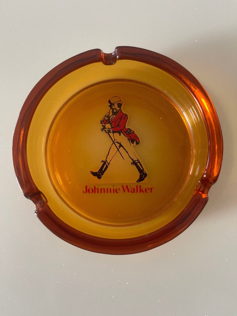 May include: Amber-colored ashtray with a circular design. The center features the Johnnie Walker logo, a man in a red coat and black boots. The brand name is printed in red below the logo. The ashtray has a smooth, glossy finish.