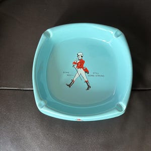 May include: A light blue square ceramic ashtray with a red and white illustration of a man in a top hat walking with a cane. The text "BORN 1620" and "STILL GOING STRONG" is printed on the ashtray.