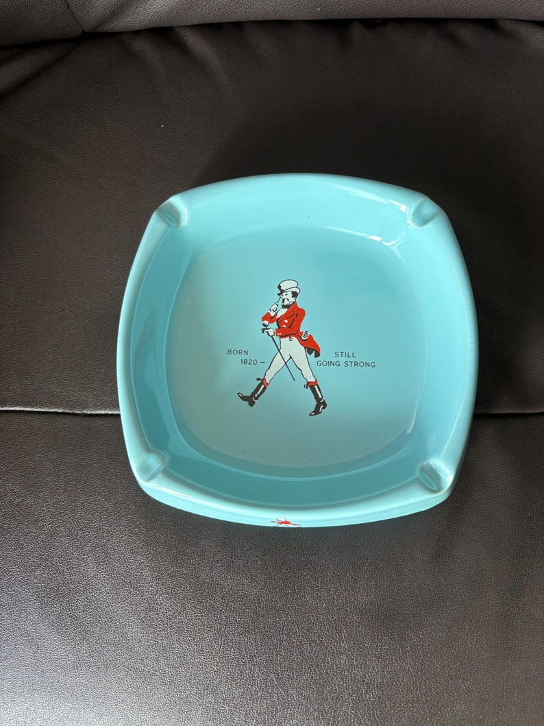 May include: A light blue square ceramic ashtray with a red and white illustration of a man in a top hat walking with a cane. The text "BORN 1620" and "STILL GOING STRONG" is printed on the ashtray.