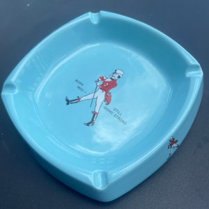 May include: A light blue ceramic ashtray with a white outline. It features a cartoon man in a red jacket and white trousers holding a cane. The text "BORN 1820 - STILL GOING STRONG" is printed on the ashtray.