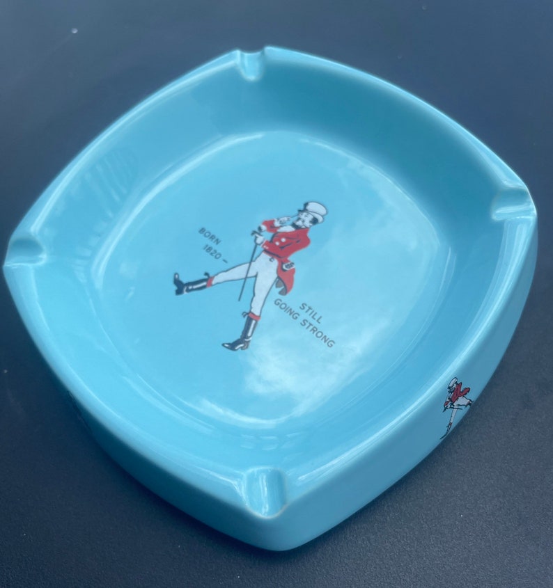 May include: A light blue ceramic ashtray with a white outline. It features a cartoon man in a red jacket and white trousers holding a cane. The text "BORN 1820 - STILL GOING STRONG" is printed on the ashtray.