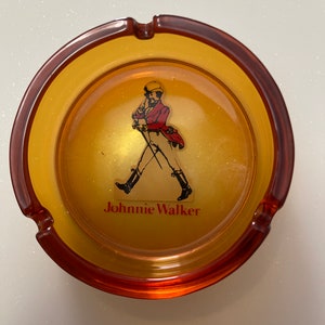 May include: Amber-colored ashtray with a brown rim. The center features the Johnnie Walker logo, a man in a red coat and black boots, with the brand name below. The ashtray has three cigarette rests.