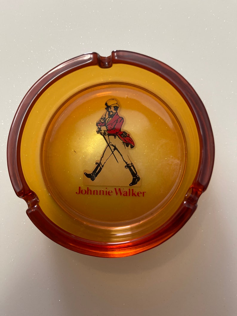 May include: Amber-colored ashtray with a brown rim. The center features the Johnnie Walker logo, a man in a red coat and black boots, with the brand name below. The ashtray has three cigarette rests.