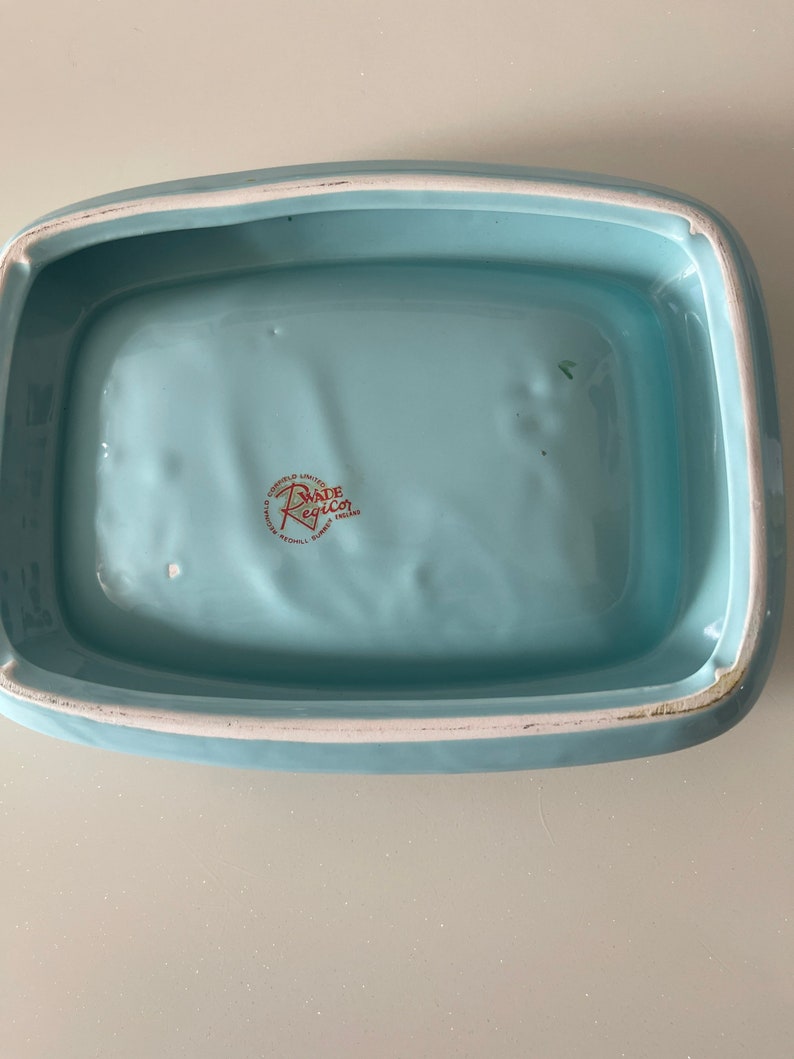 May include: A rectangular, light blue ceramic dish with rounded corners and a white rim. The interior is a solid, pale blue. The bottom of the dish has a red and white logo that reads "Wade Regicor, England".
