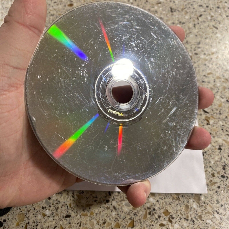 May include: A silver-colored DVD disc with rainbow-colored scratches. The disc has a black center with the text "PlayStation 2" printed on it.