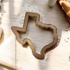 Könnte beinhalten: Ein goldfarbener Metall-Ausstecher in Form des Bundesstaates Texas. Der Ausstecher liegt auf einer Holzoberfläche mit verstreutem Mehl.