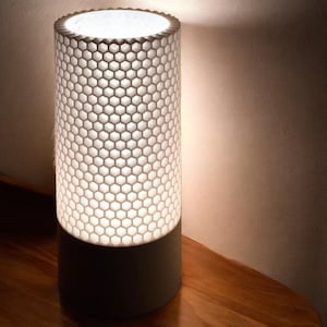 May include: White table lamp with a honeycomb-patterned shade and a white base. The lamp is turned on and illuminating the surrounding area.