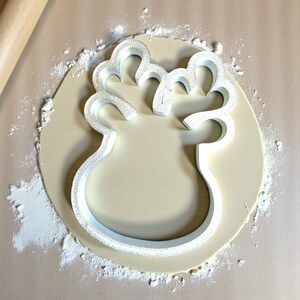 May include: A white plastic cookie cutter in the shape of a reindeer head is pressed into a circle of dough. Flour is sprinkled around the dough and the rolling pin is visible in the background. The cookie cutter has a textured surface.