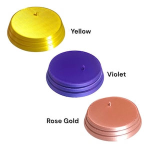 K&ouml;nnte beinhalten: Drei kreisf&ouml;rmige Pr&auml;sentationsst&auml;nder mit einem kleinen zentralen Stift, die jeweils ein gestuftes Basisdesign aufweisen. Die St&auml;nder sind in Gelb, Violett und Ros&eacute;gold gehalten, wobei die Farbnamen in schwarzem Text daneben stehen.