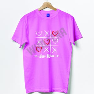 Op de afbeelding: Een lichtroze t-shirt met een tic-tac-toe ontwerp met harten en kruizen. De harten zijn rood en wit, en de tekst "Love Wins" is eronder gedrukt. Het t-shirt hangt aan een houten hanger.