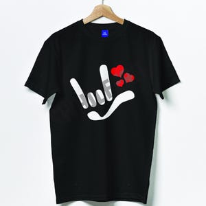 Puede incluir: Camiseta negra con un diseño de gesto de mano blanco y tres corazones rojos. La camiseta está colgada de una percha de madera. El diseño es un signo de mano de rock and roll con tres corazones rojos.