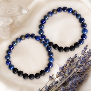 May include: Two stretch bracelets composed of round beads. One bracelet features deep blue lapis lazuli beads with gold-coloured flecks and a section of matte black beads. The second bracelet consists of similar blue lapis lazuli and black beads.