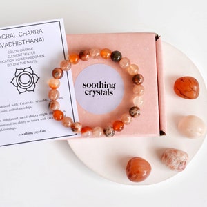 May include: A bracelet made of orange and brown carnelian beads with a pink box and carnelian stones on a white surface. The card reads "Sacral Chakra (Svadhisthana)" and "Soothing Crystals".