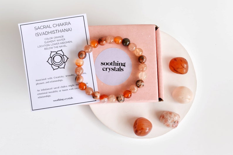 May include: A bracelet made of orange and brown carnelian beads with a pink box and carnelian stones on a white surface. The card reads "Sacral Chakra (Svadhisthana)" and "Soothing Crystals".