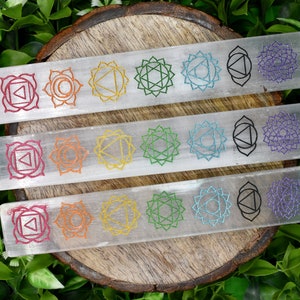 May include: Three selenite wands with the seven chakra symbols etched in different colors. The wands are on a wooden surface with green foliage in the background.