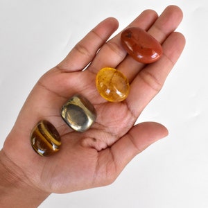 Pode incluir: Uma cole&ccedil;&atilde;o de quatro pedras polidas: uma jaspe vermelha, um olho de tigre dourado, uma citrina clara e uma pirita met&aacute;lica.