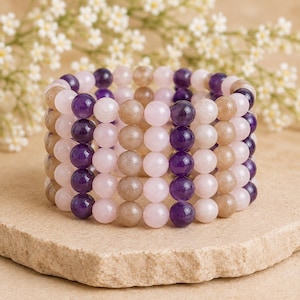 Peut inclure: Un bracelet élastique multi-rangs composé de perles lisses et rondes disposées en colonnes verticales alternées de violet profond, rose pâle et beige clair. Les perles forment une structure cylindrique avec un aspect empilé et poli.