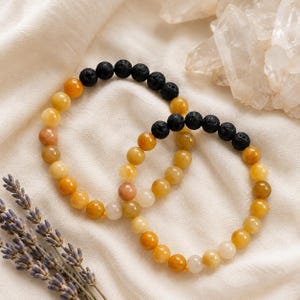 Pode incluir: Duas pulseiras de contas dispostas num padrão circular. Cada pulseira apresenta uma combinação de contas redondas e lisas de pedras preciosas amarelas e laranjas e contas de pedra de lava preta texturizadas e porosas num fio elástico.