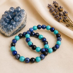 May include: Two stretch bracelets composed of round beads in shades of turquoise, deep blue, and black. The beads are arranged in a repeating pattern, creating a colorful contrast. The bracelets are displayed alongside a blue crystal cluster.