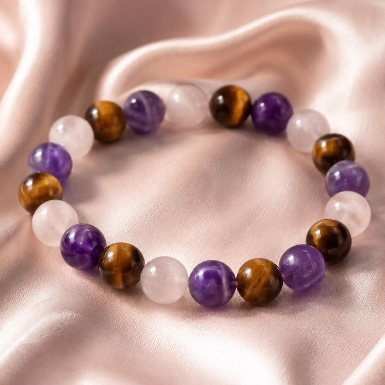 May include: A beaded bracelet featuring a repeating pattern of polished spherical beads in alternating colors of translucent light pink, deep purple, and golden brown with a chatoyant luster, arranged in a circular shape on a smooth surface.
